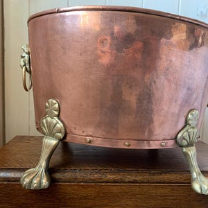 May include: A copper-colored metal tub with brass lion's head handles and brass claw feet. The tub has a rounded shape and is likely used for decorative or functional purposes. The metal has a polished finish.