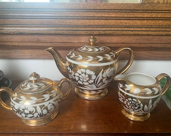 May include: A gold tea set with white floral designs. The set includes a teapot, creamer, and sugar bowl.