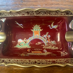 Könnte beinhalten: Ein Aschenbecher aus roter Keramik mit goldenen Verzierungen und einem chinesisch inspirierten Design. Der Aschenbecher zeigt eine Pagode, eine Brücke und zwei Vögel. Das Design ist in Gold und Weiß auf rotem Untergrund gemalt.