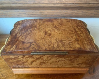 May include: A wooden box with a hinged lid and a brass hinge. The box is made of a light brown wood with a dark brown wood grain pattern. The box has a decorative edge around the top and bottom.