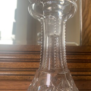 May include: A clear glass decanter with a stopper and a cut glass design. The decanter is sitting on a wooden surface.