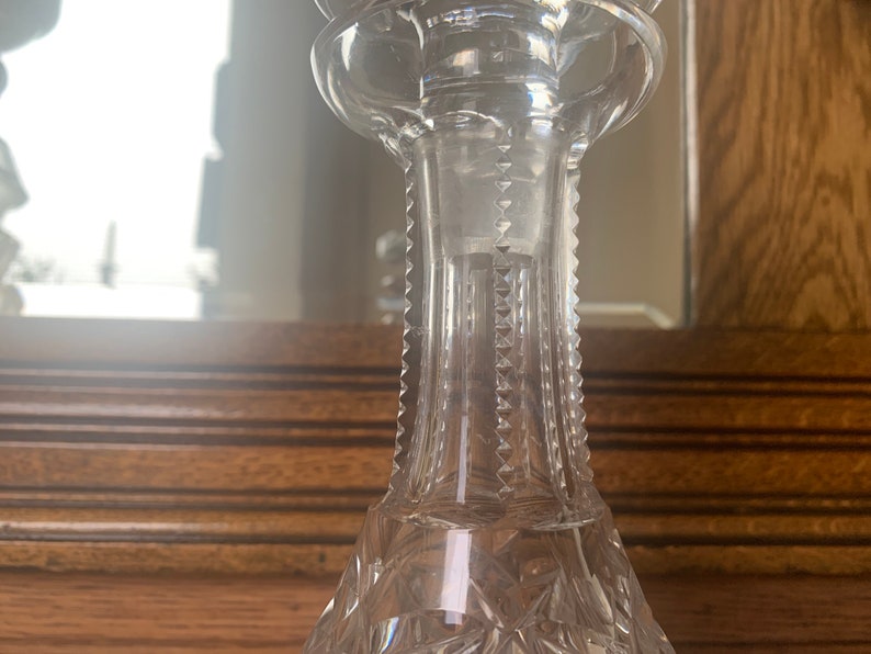May include: A clear glass decanter with a stopper and a cut glass design. The decanter is sitting on a wooden surface.
