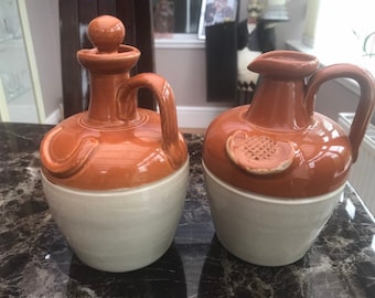 Puede incluir: Dos jarras de cerámica con esmalte marrón y blanco. Las jarras tienen un cuerpo redondeado y un pico. Una jarra tiene una tapa.