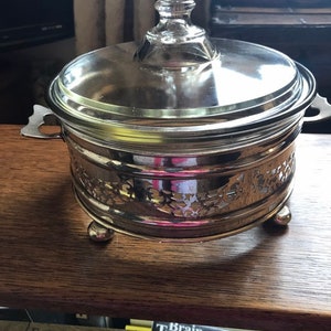 Könnte beinhalten: Silberne Servierplatte mit Glasdeckel und verziertem Design. Die Platte hat drei Füße und zwei Griffe. Die Platte steht auf einer Holzoberfläche.