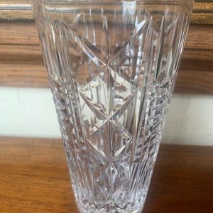 Puede incluir: Un jarrón de vidrio transparente con un diseño de cristal tallado. El jarrón tiene una forma alta y cilíndrica y una boca ancha.