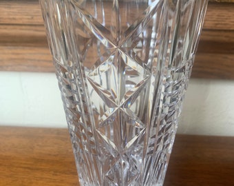 May include: A clear glass vase with a cut crystal design. The vase has a tall, cylindrical shape and a wide mouth.