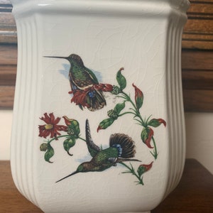 Może przedstawiać: Biały ceramiczny wazon z sześciokątną podstawą i lekko rozszerzaną górą. Wazon przedstawia szczegółową ilustrację dwóch kolibrów z czerwonymi kwiatami i zielonymi liśćmi. Wazon ma spękaną powierzchnię.