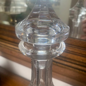 May include: A clear glass decanter with a detailed, faceted design. The top of the decanter has a decorative, multi-layered, circular stopper.