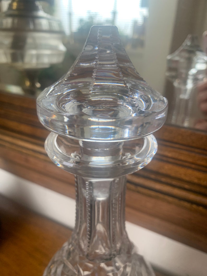 May include: A clear glass decanter with a detailed, faceted design. The top of the decanter has a decorative, multi-layered, circular stopper.