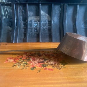 Pode incluir: Uma concha de cobre com uma alça longa, apoiada em uma superfície de madeira com um design floral. A concha está ligeiramente desbotada e tem uma tigela arredondada.