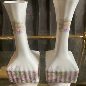 Op de afbeelding: Twee witte porseleinen vazen met een bloemenmotief. De vazen hebben een geribbeld ontwerp en zijn versierd met roze en paarse bloemen.