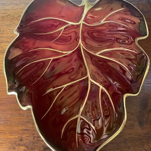 Könnte beinhalten: Eine rote, blattförmige Servierplatte mit goldenen Verzierungen. Die Platte ist aus Keramik und hat eine glänzende Oberfläche.