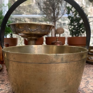 Puede incluir: Un cubo de latón con un asa negra. El cubo es redondo y tiene una superficie lisa. Está sentado en una encimera de granito.