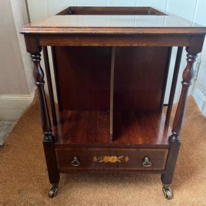 Puede incluir: Un carrito de té de madera oscura con una parte superior rectangular y un estante inferior. El carrito tiene un cajón con incrustaciones florales y está sostenido por patas torneadas y pequeñas ruedas. La madera tiene un tono rico y cálido.