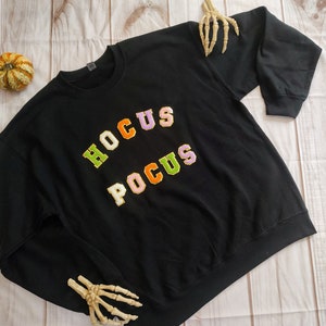 May include: Black sweatshirt with the words "Hocus Pocus" in colorful letters. The letters are in a varsity style font.
