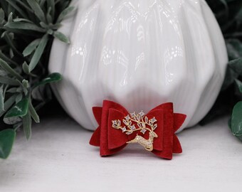 May include: A red velvet bow with a gold-colored deer and floral embellishments. The bow is set against a white background with green foliage, creating a festive and elegant aesthetic.