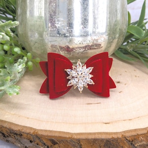 May include: A red velvet bow with a sparkling crystal snowflake center. The bow is set on a wooden slice, with greenery and a silver glass container in the background. This decorative bow is suitable for various crafts and holiday decorations.