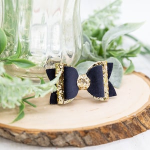 K&ouml;nnte beinhalten: Eine schwarze Haarklammer mit goldener Glitzer Schleife. Die Schleife ist aus schwarzem Stoff mit goldenen Glitzer Akzenten. Die Klammer ist an der R&uuml;ckseite der Schleife befestigt.