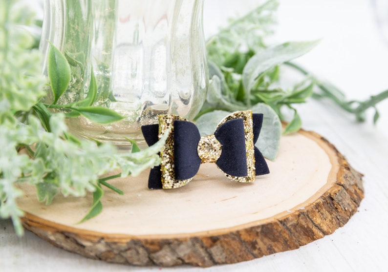 K&ouml;nnte beinhalten: Eine schwarze Haarklammer mit goldener Glitzer Schleife. Die Schleife ist aus schwarzem Stoff mit goldenen Glitzer Akzenten. Die Klammer ist an der R&uuml;ckseite der Schleife befestigt.
