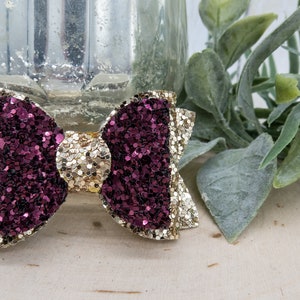 May include: A gold and burgundy glitter bow hair clip. The bow is made of two layers of glitter fabric, one gold and one burgundy. The bow is attached to a metal clip.