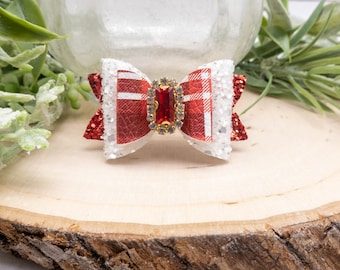 May include: A decorative hair bow featuring a red and white plaid pattern with glitter accents. The bow is adorned with a rectangular, red gemstone centerpiece surrounded by smaller, clear stones. The bow is set on a wooden surface.