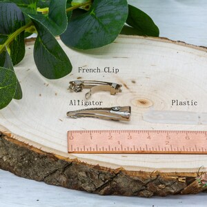 K&ouml;nnte beinhalten: Drei Arten von Haarklammern: eine silberne Alligator-Klammer, eine silberne franz&ouml;sische Klammer und eine transparente Plastikklammer. Die Klammern befinden sich auf einer Holzoberfl&auml;che mit einem Lineal als Gr&ouml;&szlig;enreferenz. Der Text "French Clip", "Alligator" und "Plastic" ist auf der Oberfl&auml;che geschrieben.