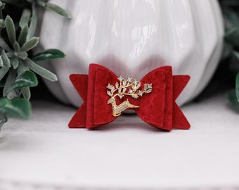 May include: A red velvet bow with a gold and rhinestone deer design. The bow is centered in the image, with a white background and green foliage in the background.