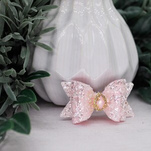 May include: A pink glitter bow with a gold-colored jewel in the center. The bow is set against a white background with green foliage. The bow is likely for a pet or a hair accessory.