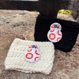 May include: Two rectangular crochet cup sleeves, one in solid black and one in off-white, each featuring a centered embroidered patch of a white, orange, and gray spherical robot character with a circular head and a round body design.