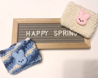Op de afbeelding: Twee handgemaakte gehaakte bekerhoezen, één crèmekleurig met een roze konijnenapplicatie en één blauw en wit met een blauwe konijnenapplicatie. De zin "HAPPY SPRING" wordt weergegeven op een grijs letterbord.