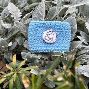 K&ouml;nnte beinhalten: Eine hellblaue, handgestrickte Kaffeebecherh&uuml;lle mit einem wei&szlig;-schwarzen, gestickten Aufn&auml;her. Der Aufn&auml;her zeigt eine Strichzeichnung des Profils einer Frau. Die H&uuml;lle wird vor einem Hintergrund aus gr&uuml;nem und grauem Laubwerk pr&auml;sentiert.