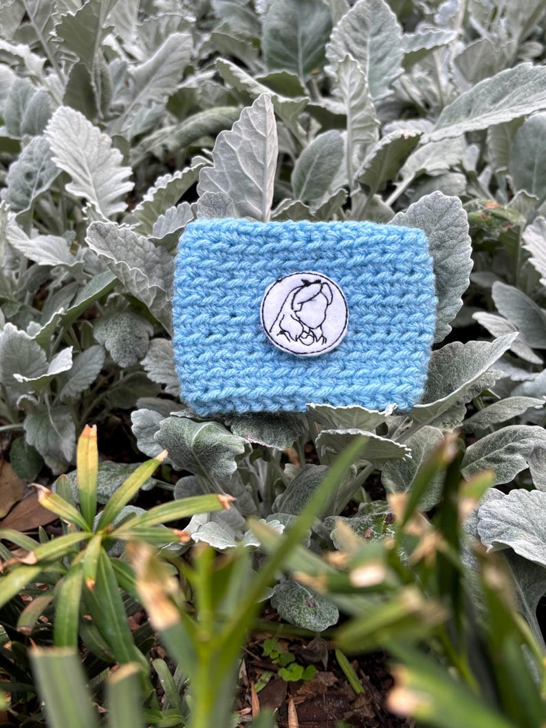 K&ouml;nnte beinhalten: Eine hellblaue, handgestrickte Kaffeebecherh&uuml;lle mit einem wei&szlig;-schwarzen, gestickten Aufn&auml;her. Der Aufn&auml;her zeigt eine Strichzeichnung des Profils einer Frau. Die H&uuml;lle wird vor einem Hintergrund aus gr&uuml;nem und grauem Laubwerk pr&auml;sentiert.