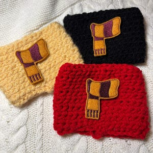 May include: Three handmade crochet cup sleeves in yellow, black, and red. Each sleeve features a sewn-on patch of a scarf in gold, purple, and yellow. The sleeves are displayed on a white, textured surface.