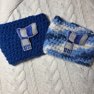 May include: Two blue crochet cup sleeves with a scarf applique. One sleeve is solid blue, the other is blue and white striped. The sleeves are on a white cable knit background.