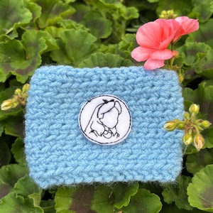 K&ouml;nnte beinhalten: Eine hellblaue geh&auml;kelte Kaffeebecherh&uuml;lle mit einem wei&szlig;en runden Aufn&auml;her mit einer schwarz-wei&szlig;en Illustration des Profils eines M&auml;dchens. Die H&uuml;lle ist von gr&uuml;nem Laub und einer rosa Blume umgeben.