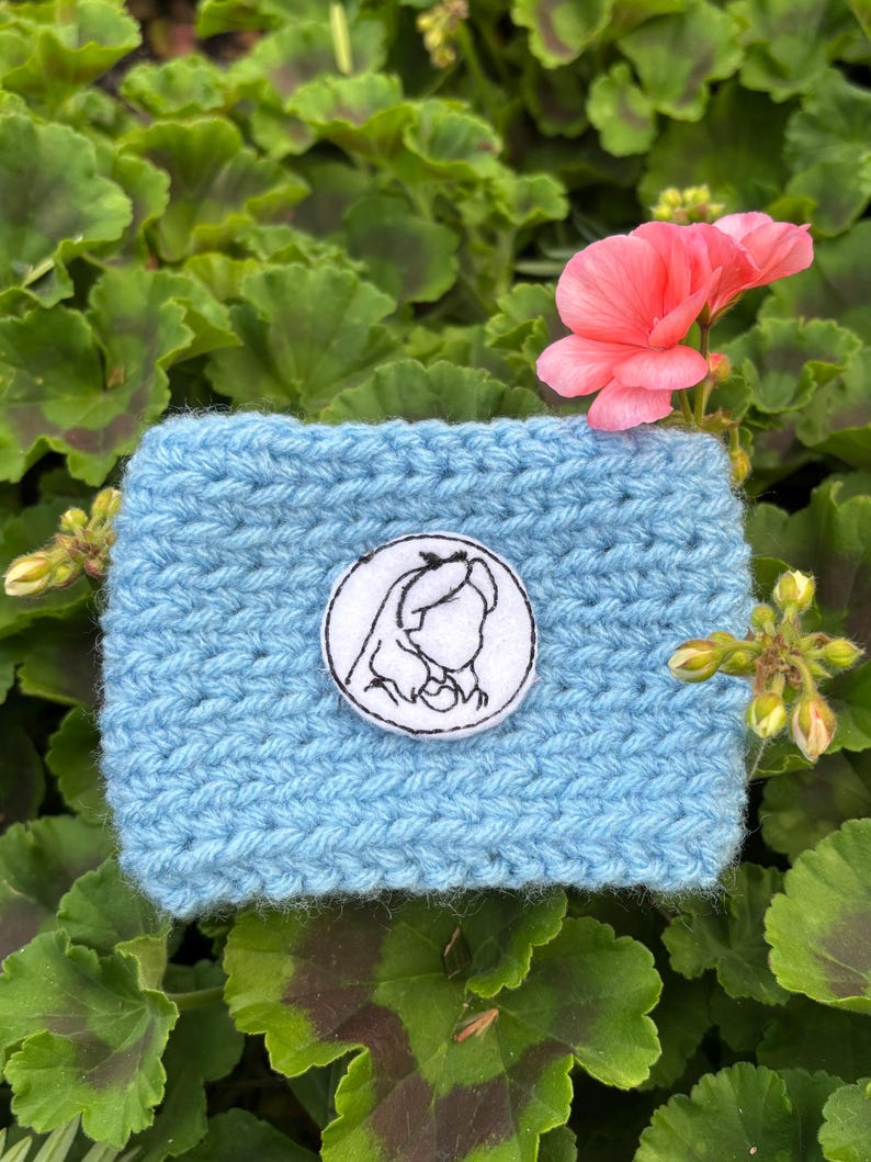 K&ouml;nnte beinhalten: Eine hellblaue geh&auml;kelte Kaffeebecherh&uuml;lle mit einem wei&szlig;en runden Aufn&auml;her mit einer schwarz-wei&szlig;en Illustration des Profils eines M&auml;dchens. Die H&uuml;lle ist von gr&uuml;nem Laub und einer rosa Blume umgeben.