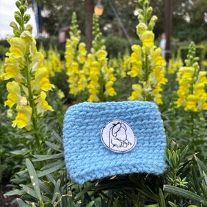 K&ouml;nnte beinhalten: Eine hellblaue, gestrickte Kaffeebecherh&uuml;lle mit einem wei&szlig;-schwarzen, gestickten Aufn&auml;her mit einem Charakterdesign. Die H&uuml;lle ist von gr&uuml;nem Laub und gelben Blumen umgeben.