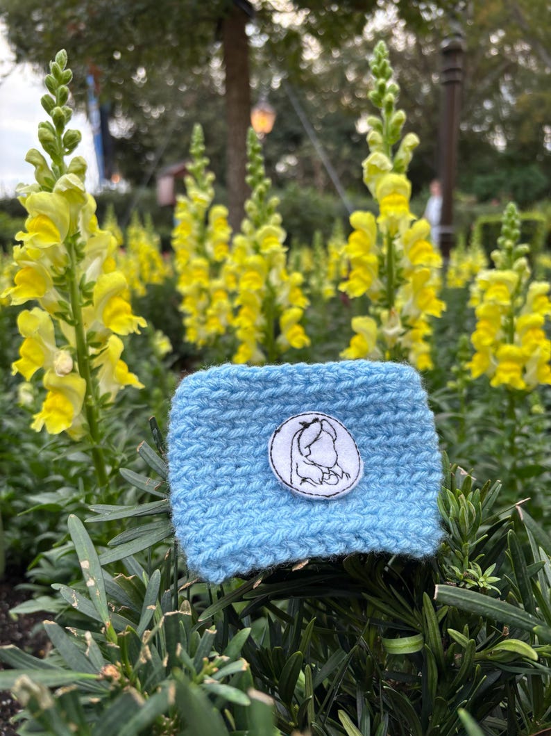 K&ouml;nnte beinhalten: Eine hellblaue, gestrickte Kaffeebecherh&uuml;lle mit einem wei&szlig;-schwarzen, gestickten Aufn&auml;her mit einem Charakterdesign. Die H&uuml;lle ist von gr&uuml;nem Laub und gelben Blumen umgeben.