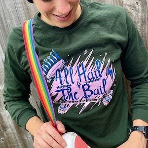 May include: Navy blue beanie with roller skate embroidery and a dark green long-sleeve shirt with the text "All Hail The Bail". A rainbow strap is attached to a pair of pink roller skates.