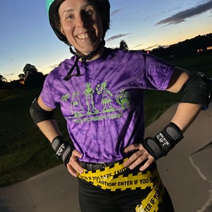 Puede incluir: Una mujer con una camiseta de color morado tie-dye con un gráfico de temática de Halloween que dice "The Scream Team". Lleva un casco turquesa, pantalones negros y cinta amarilla de precaución alrededor de la cintura.