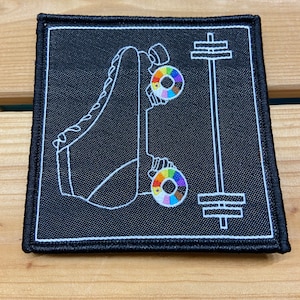 Op de afbeelding: Een geborduurde patch in zwart en wit met een rolstoel met een regenboogkleurig wiel en een halterstang. De patch heeft een zwarte achtergrond en witte stiksels.