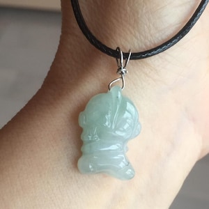May include: A pale green jade pendant necklace with a carved figure. The pendant hangs from a black cord. The jade is translucent, and the figure has visible facial details. The necklace is displayed on a wrist.