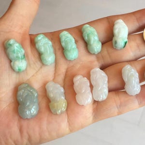 Könnte beinhalten: Eine Sammlung kleiner, geschnitzter Jadefiguren in Grün-, Weiß- und Grautönen. Die Figuren werden in einer Hand gehalten, wobei auch zwei Münzen zu sehen sind. Die Jadestücke haben ein glattes, poliertes Aussehen.