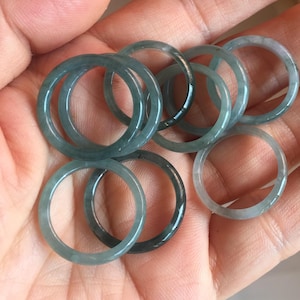 May include: A collection of jade rings in varying shades of green and gray. The rings are circular and appear to be made of a translucent stone. The rings are held in a person's open palm, showcasing their natural color and design.