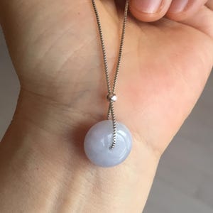 May include: A silver necklace with a circular, pale lavender pendant. The pendant is smooth and polished, hanging from a fine chain. The necklace is held in a hand, highlighting the jewellery's design and colour.