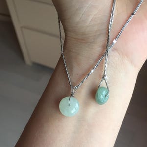 May include: Two silver necklaces with jade pendants. One necklace has a round, pale green jade pendant, the other a teardrop-shaped, pale green jade pendant. The necklaces are displayed on a person's arm.