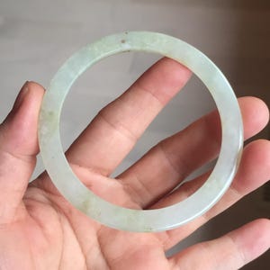 May include: A pale green jade bangle bracelet. The circular bracelet has a smooth, polished surface. The jade has subtle variations in color, with areas of lighter and darker green. The bracelet is held in a hand.