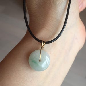 May include: A circular, pale green jade pendant with a gold-coloured wire loop, hanging from a black cord necklace. The jade has subtle green veining. The necklace is displayed against a skin-toned background.