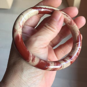 May include: A polished bangle bracelet featuring a smooth, rounded surface with a marbled pattern of deep red and creamy beige tones. The circular accessory displays natural colour variations and a glossy finish throughout its entire circumference.