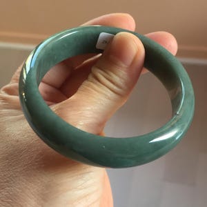 May include: A jade green bangle bracelet. The smooth, polished surface reflects light, showcasing the natural variations in colour. The bracelet is a solid, circular design, and is held in a hand.